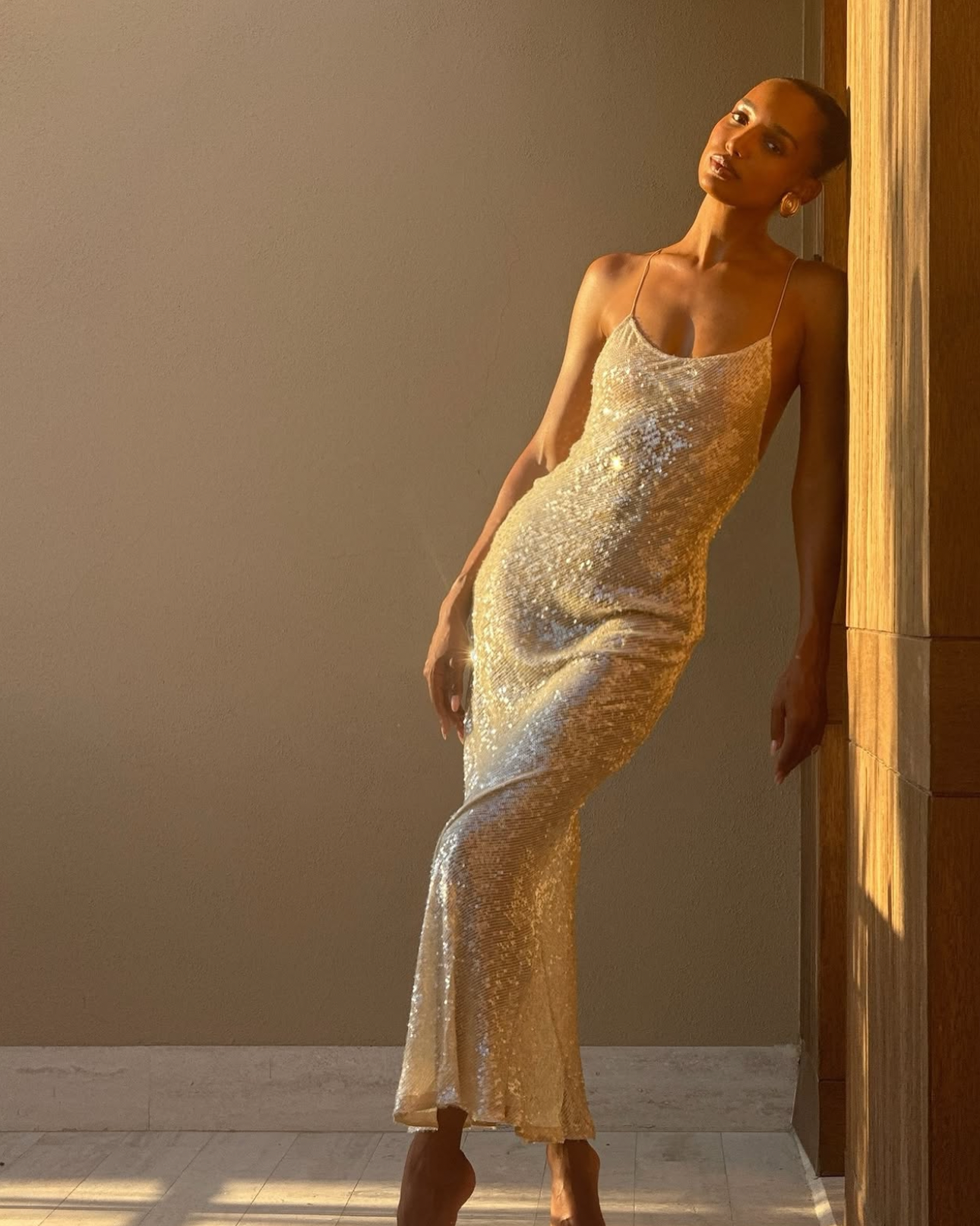 A woman wearing a sleeveless, fitted champagne-gold sequined dress posing in a sunlit indoor setting. The photo appears to be from a social media post, with the caption text partially visible and a large gray overlay obscuring the center.