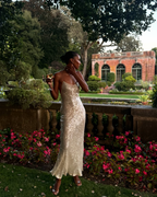 A woman wearing a sleeveless champagne-gold sequined evening gown stands outdoors in a garden setting with pink flowers and a brick building in the background, holding a drink. This appears to be a social media post screenshot rather than a clean product listing photo.