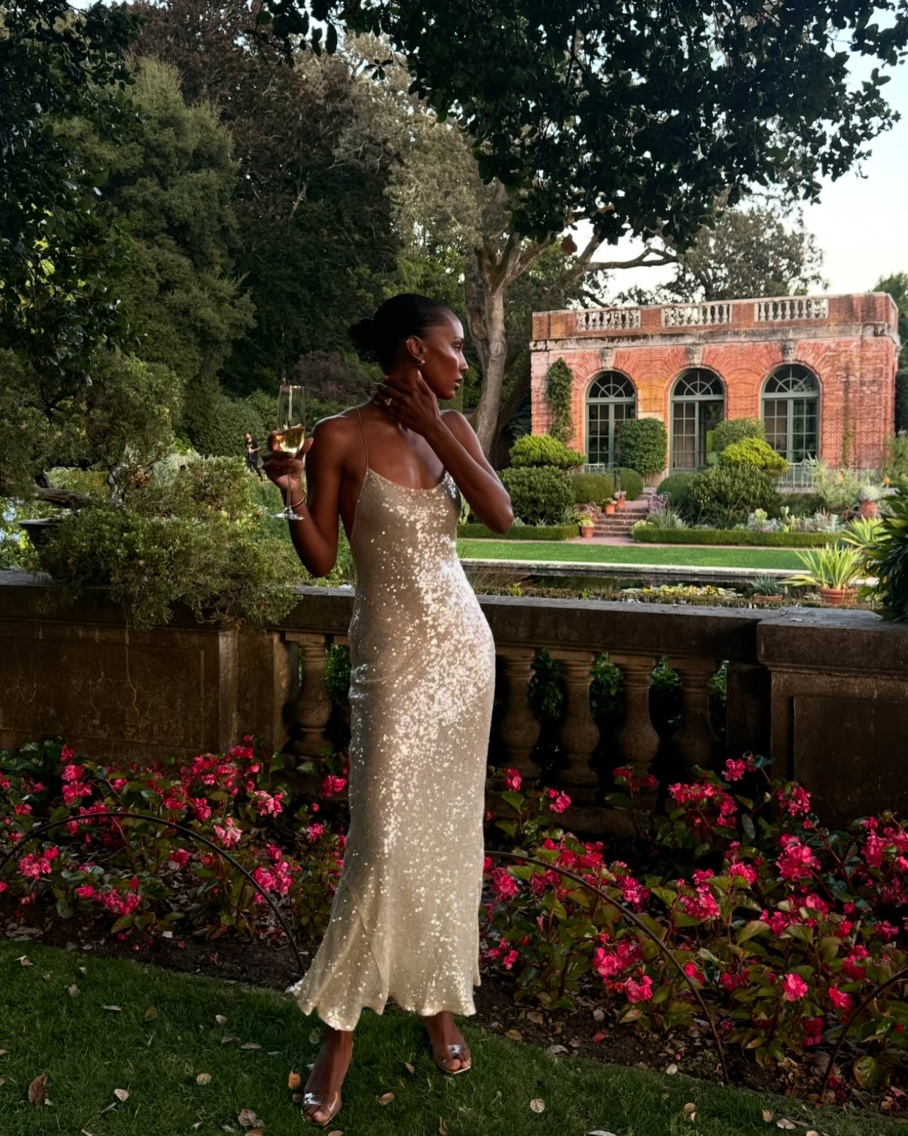 A woman wearing a sleeveless champagne-gold sequined evening gown stands outdoors in a garden setting with pink flowers and a brick building in the background, holding a drink. This appears to be a social media post screenshot rather than a clean product listing photo.