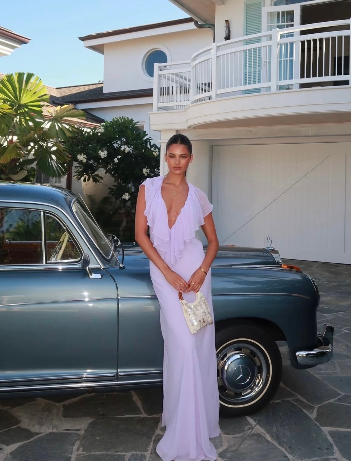 A woman wearing a lavender sleeveless ruffled maxi dress stands in front of a vintage blue car on a stone driveway in front of a white house.