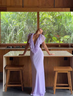 A woman wearing a lavender purple ruffled maxi dress stands with her back to the camera, leaning on a kitchen island with wooden stools in front of large windows showing lush green foliage outside.