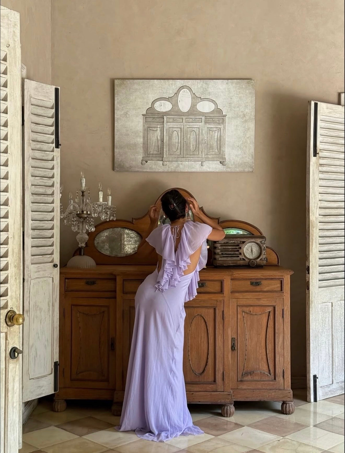 A woman stands with her back to the camera in a lavender purple sleeveless maxi dress, posing in front of a vintage wooden sideboard. The room features beige walls, a framed cabinet painting, and white shutters on the sides.