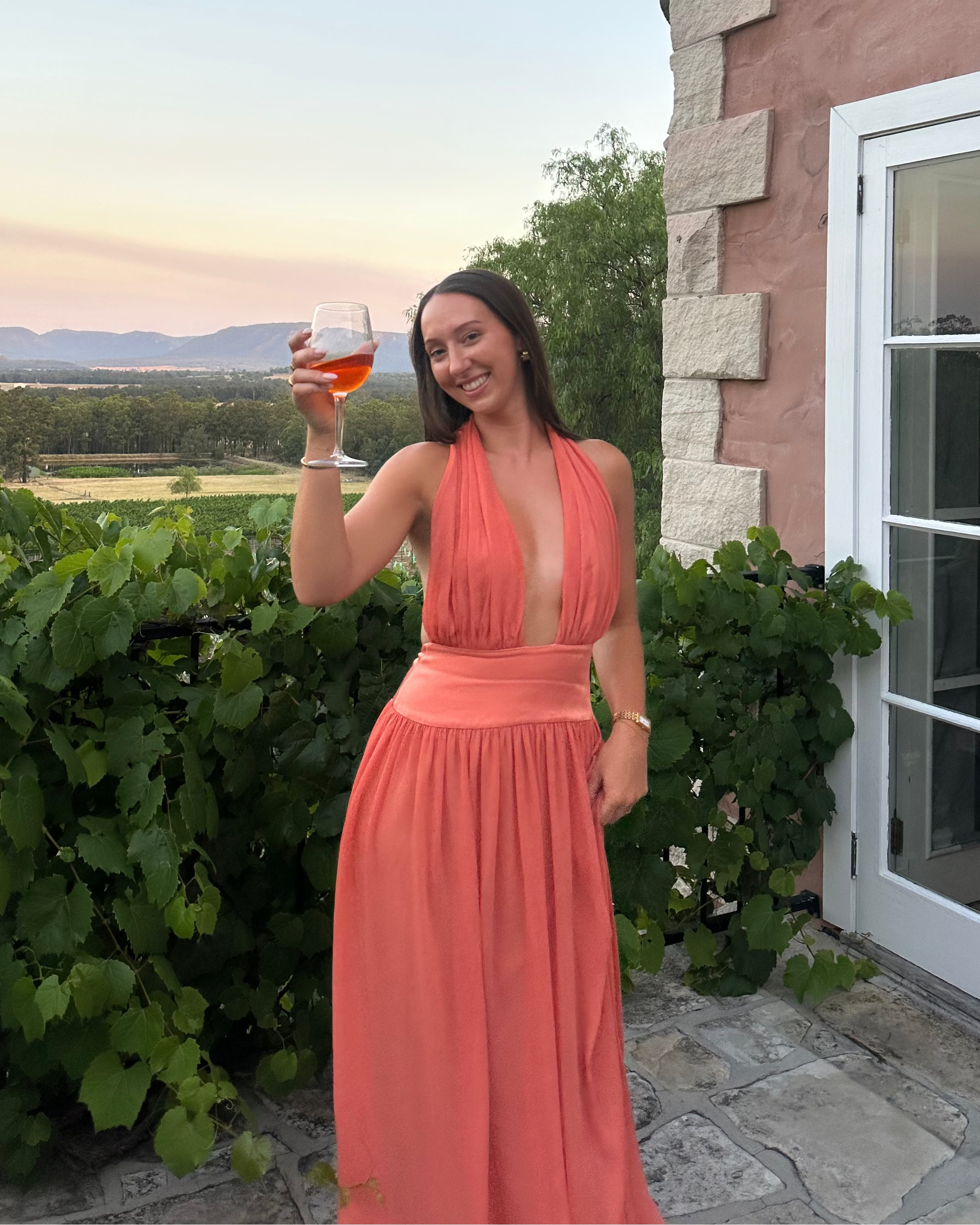 Woman in a coral dress holding a glass of wine outside a stone building with vineyard view.